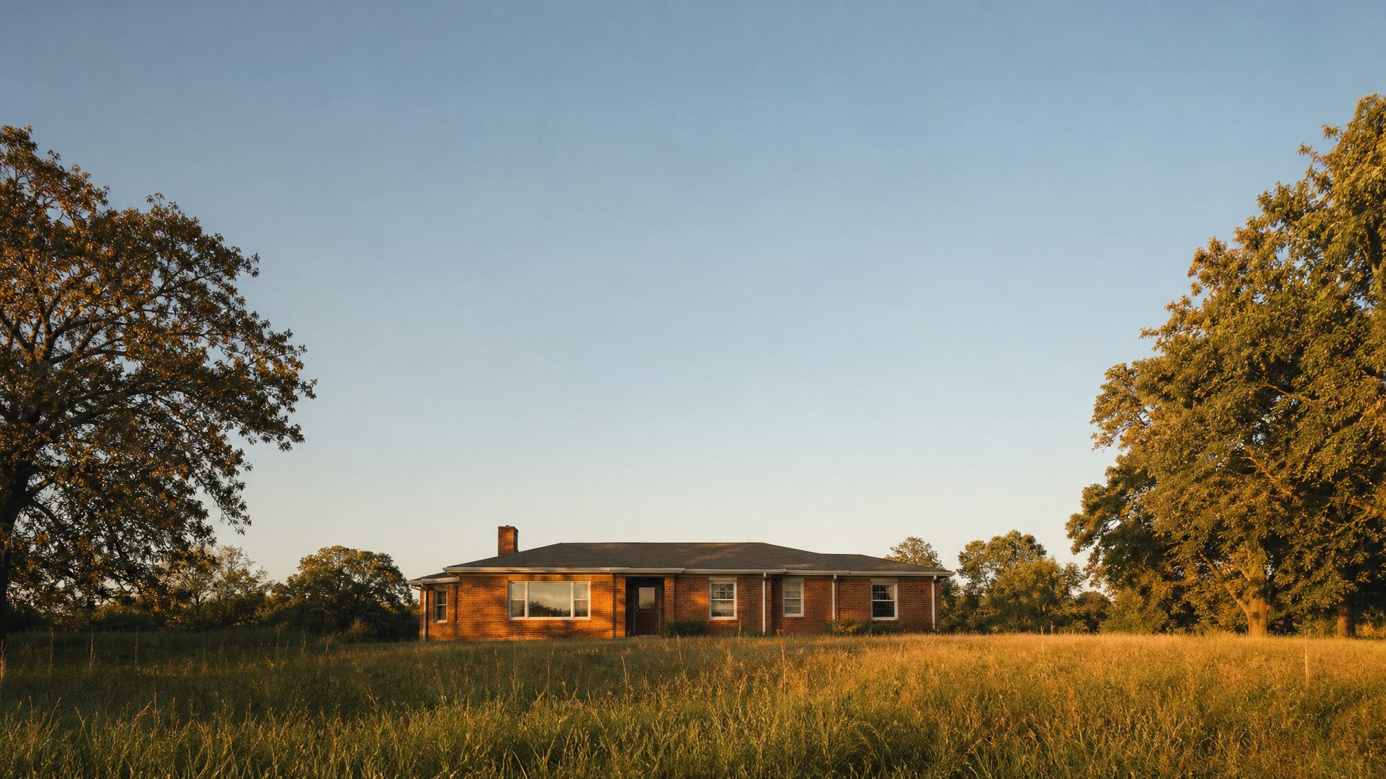 A modest single story brick house sitting in an open grassy lot at golden hour, with a wide clear evening sky above it.
